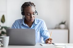 a woman sitting at a desk with a laptop and headphones on, writing in a notebook