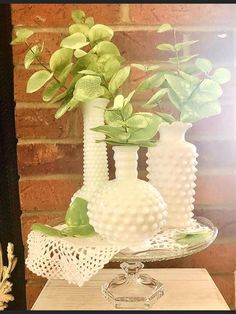 three white vases with green plants in them sitting on a small table next to a brick wall