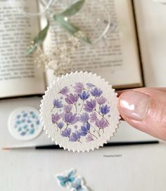 a hand holding a sticker with purple flowers on it next to an open book