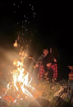 two people sitting around a campfire with their arms in the air and one person standing next to it