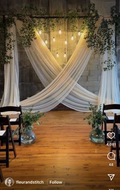 an indoor wedding ceremony with white drapes and greenery draped over the back of chairs