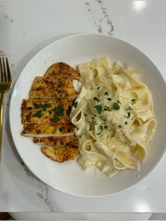 a white plate topped with pasta and meat next to a fork on a marble table