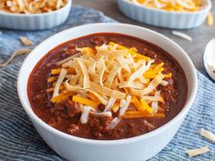 two bowls filled with chili and cheese on top of a blue towel next to spoons