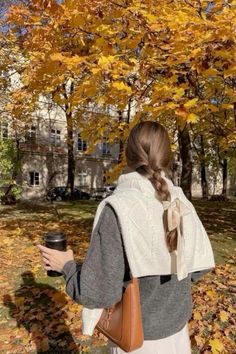 a woman holding a cup in her hand while standing under a tree with yellow leaves