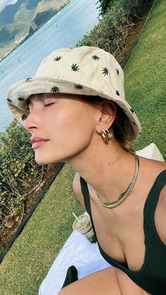 a woman wearing a hat sitting on top of a grass covered field next to a body of water