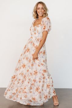 a woman wearing a white and orange floral print dress, smiling at the camera with her hands in her pockets