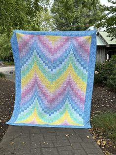 a blue, yellow and pink quilt sitting on top of a brick walkway next to trees