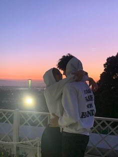 two people in hoodies are standing on a balcony at night with the sun setting behind them