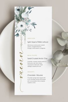 a menu card with white flowers and greenery on it, next to a plate