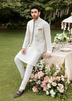 a man sitting on top of a table with flowers in front of him wearing a white suit and tie