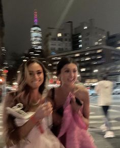 two women in dresses are posing for the camera on a city street at night with skyscrapers in the background