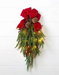 a christmas wreath with pine cones and red ribbon hanging on a white wooden wall, decorated with lights