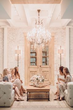 a group of women sitting around a table in a living room with chandelier