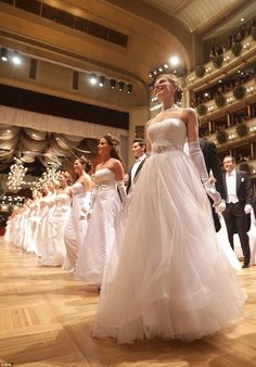 a group of women in white dresses walking down a wooden floor with men in suits and tuxedos behind them