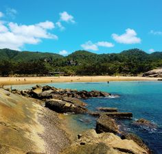 people are swimming in the ocean near some rocks and sand on a sunny day with blue skies