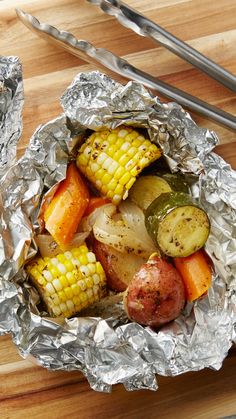 a wooden table topped with aluminum foil covered in vegetables and corn on the cob