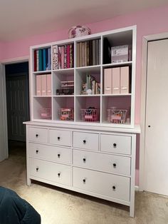 a white dresser with lots of books on top of it in a pink walled room