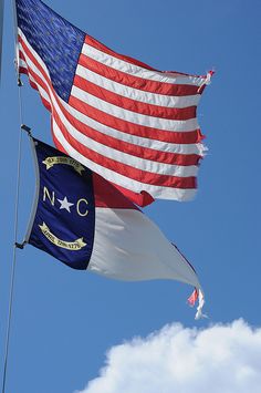 two american and north carolina flags flying side by side against a blue sky with white clouds