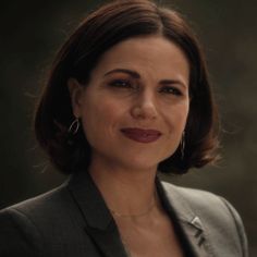 a woman with short hair wearing a gray suit and smiling at the camera while standing in front of a dark background