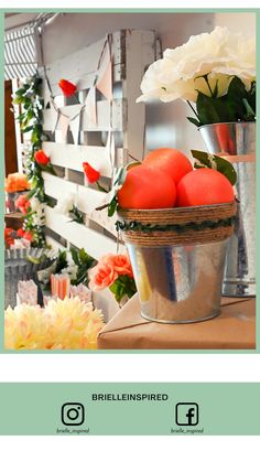 there is a bucket full of oranges on the table with flowers in front of it