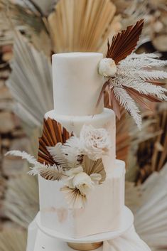 a three tiered white wedding cake with flowers and feathers on the top is surrounded by palm leaves