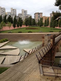 an old bridge over a small pond in the middle of a park with buildings in the background