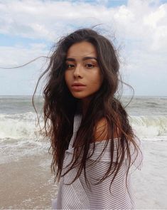 a woman standing on top of a beach next to the ocean with her hair blowing in the wind