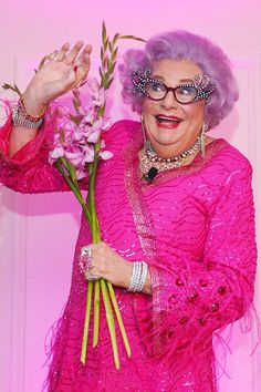 an older woman in pink holding flowers and posing for the camera with her hands up