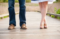 a man and woman walking on a wooden bridge holding each other's hands, with grass in the background