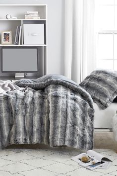 a bed with grey and white fur on it in front of a computer desk, bookshelf and window