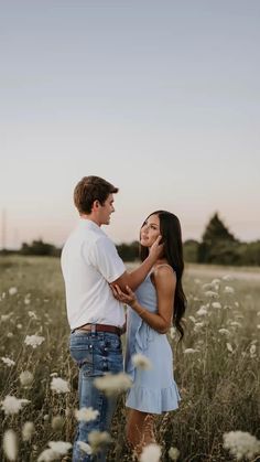 a man and woman standing in a field with tall grass at sunset, looking into each other's eyes