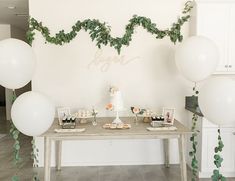 a table with white balloons and greenery on the wall, along with desserts