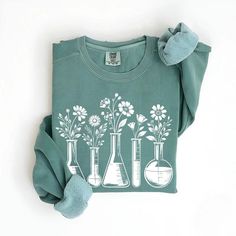 Embrace the cozy side of science with our Sleek Beaker Botanical Sweatshirt. This unique pullover showcases a stunning design of delicate flowers emerging from various laboratory beakers and flasks, perfectly capturing the intersection of botany and chemistry. Ideal for chilly days in the lab or classroom, this sweatshirt offers both warmth and style to science teachers, chemists, and biology enthusiasts alike. The soothing sage green color complements the intricate white floral and glassware design, creating a piece that's both sophisticated and fun. Whether you're conducting experiments, grading papers, or simply enjoying a casual day out, this sweatshirt is sure to spark curiosity and admiration from fellow science lovers. It's the perfect way to wear your passion for science and nature Biology Lab, Biology Labs, Glassware Design, Lab Technician, Sage Green Color, Beakers, Floral Sweater, Color Complement, Science Teacher