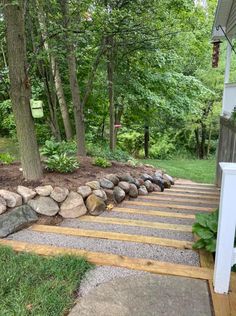 a garden with rocks and wood steps leading up to the tree lined area in front of the house