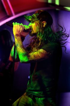 a man with long hair holding a microphone in front of purple and green lights at a concert