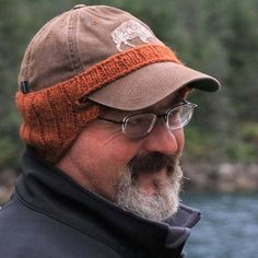 a bearded man wearing a hat and glasses next to a body of water with trees in the background