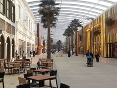 people are walking through an indoor mall with tables and chairs on the ground, palm trees in the background