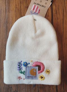 a white beanie with flowers on it sitting on top of a wooden table next to a tag