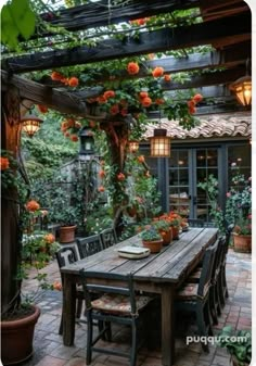 an outdoor dining table surrounded by potted plants and hanging orange flowers on the pergolated roof