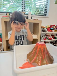 a young boy wearing glasses looking at a volcano in a play room with toys on the floor