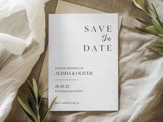 a wedding save the date card on top of a bed with white sheets and greenery