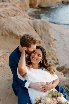 a man and woman are sitting on the beach with their arms around each other as they hug