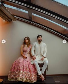 a man and woman sitting next to each other in front of a white wall wearing wedding outfits