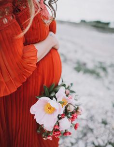 a pregnant woman in an orange dress holding a bouquet of white and red flowers on her belly