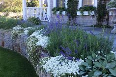 a stone wall with flowers and plants growing on it in front of a house next to a lawn