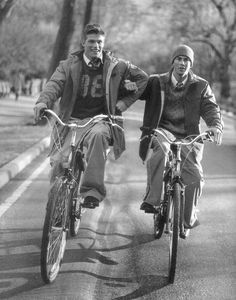 two men riding bikes down the street in front of each other, one is wearing a jacket and the other has a hat