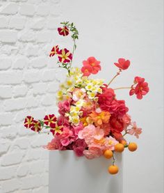 a white vase filled with lots of colorful flowers next to a brick wall and oranges