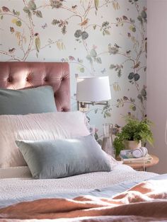 a bed with pillows and blankets on it next to a night stand in front of a floral wallpaper