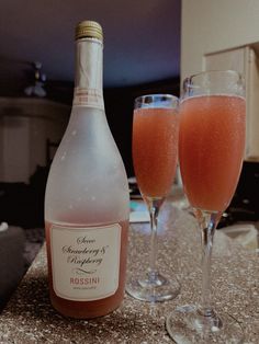 two wine glasses filled with pink liquid next to a bottle of sparkling champagne on a counter