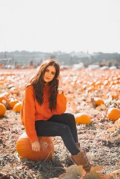 a woman in an orange sweater is sitting on a pumpkin patch with her legs crossed